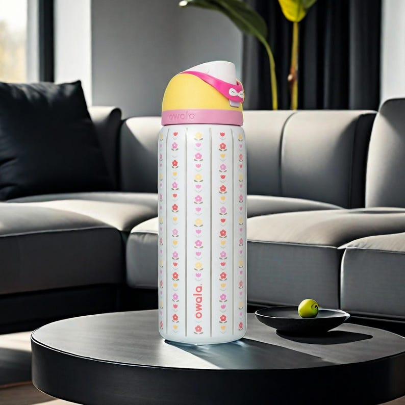 Puede incluir: Una botella de agua Owala blanca con la parte superior amarilla y detalles en rosa. La botella presenta un estampado floral con rayas verticales. La botella est&aacute; sobre una mesa negra con un cuenco peque&ntilde;o y una fruta verde.