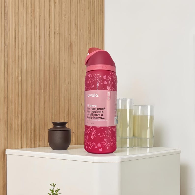 Puede incluir: Una botella de agua Owala roja con un estampado floral. La botella tiene una pajita incorporada y una tapa abatible. El texto de la botella dice: "Hola. Soy a prueba de fugas. Estoy aislado. Y tengo una pajita incorporada."