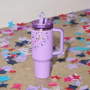 Peut inclure: Un gobelet Stanley lavande avec une poign&eacute;e et une paille. Le gobelet pr&eacute;sente un motif d'&eacute;toiles et le logo Stanley. Le couvercle est d'une nuance de violet plus fonc&eacute;e. Le gobelet est entour&eacute; de confettis en forme d'&eacute;toiles.
