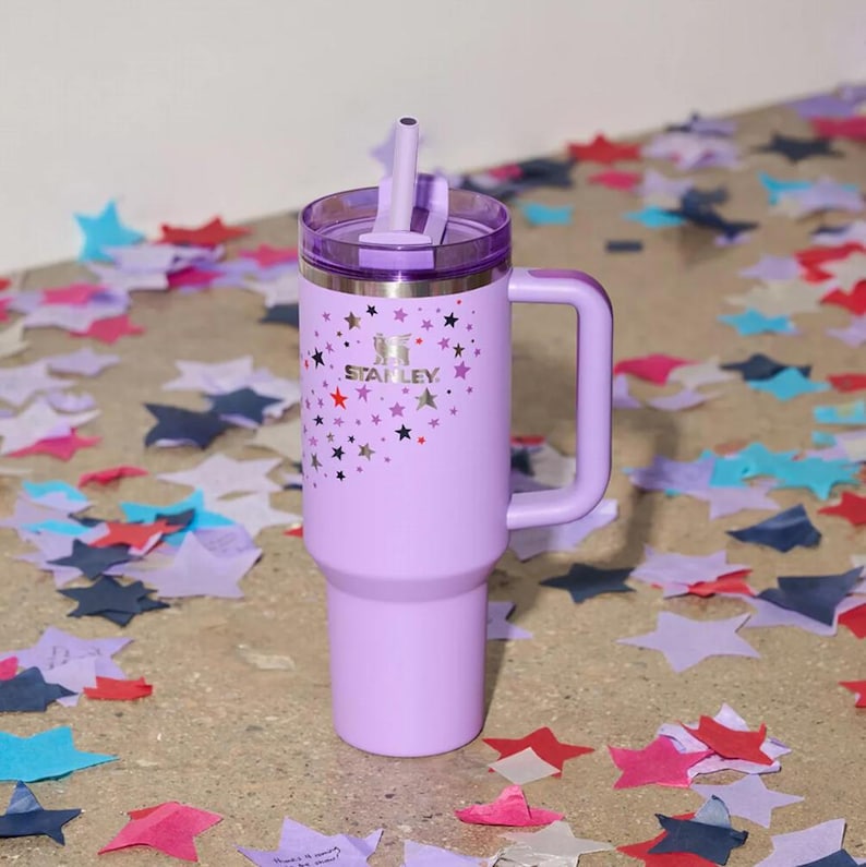 Peut inclure: Un gobelet Stanley lavande avec une poign&eacute;e et une paille. Le gobelet pr&eacute;sente un motif d'&eacute;toiles et le logo Stanley. Le couvercle est d'une nuance de violet plus fonc&eacute;e. Le gobelet est entour&eacute; de confettis en forme d'&eacute;toiles.