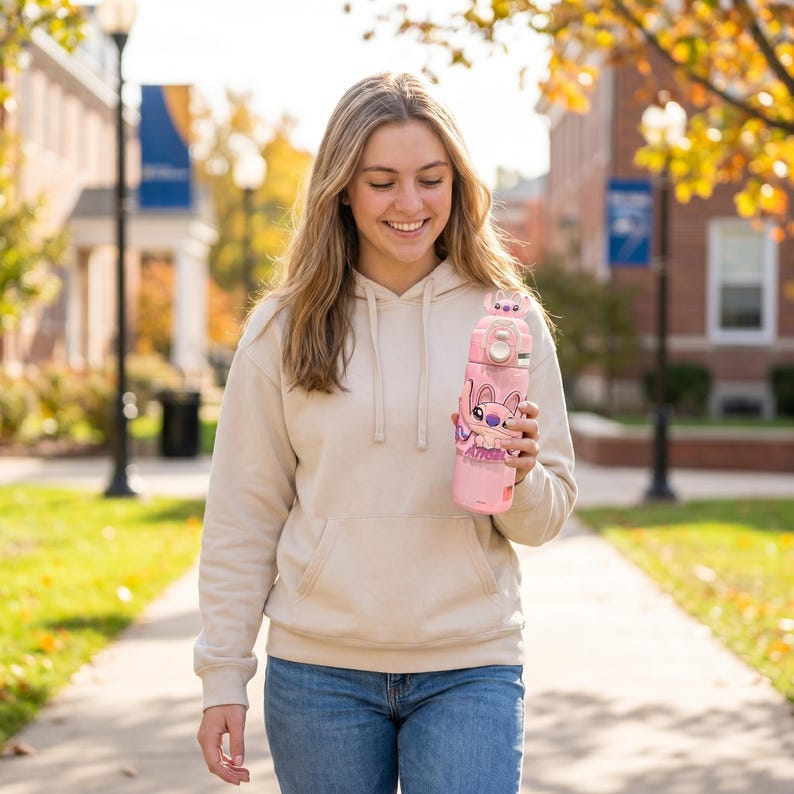 May include: A pink water bottle with a cartoon character design. The bottle has a light pink body with a darker pink top and a character illustration. The bottle is held by a person wearing a beige hoodie and blue jeans.