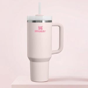 Op de afbeelding: Een lichtroze Stanley-beker met een handvat en een wit deksel. De beker heeft een roze logo en een wit rietje. De beker is ontworpen om een grote hoeveelheid vloeistof te bevatten.