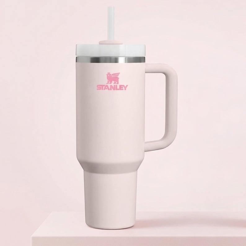 Op de afbeelding: Een lichtroze Stanley-beker met een handvat en een wit deksel. De beker heeft een roze logo en een wit rietje. De beker is ontworpen om een grote hoeveelheid vloeistof te bevatten.