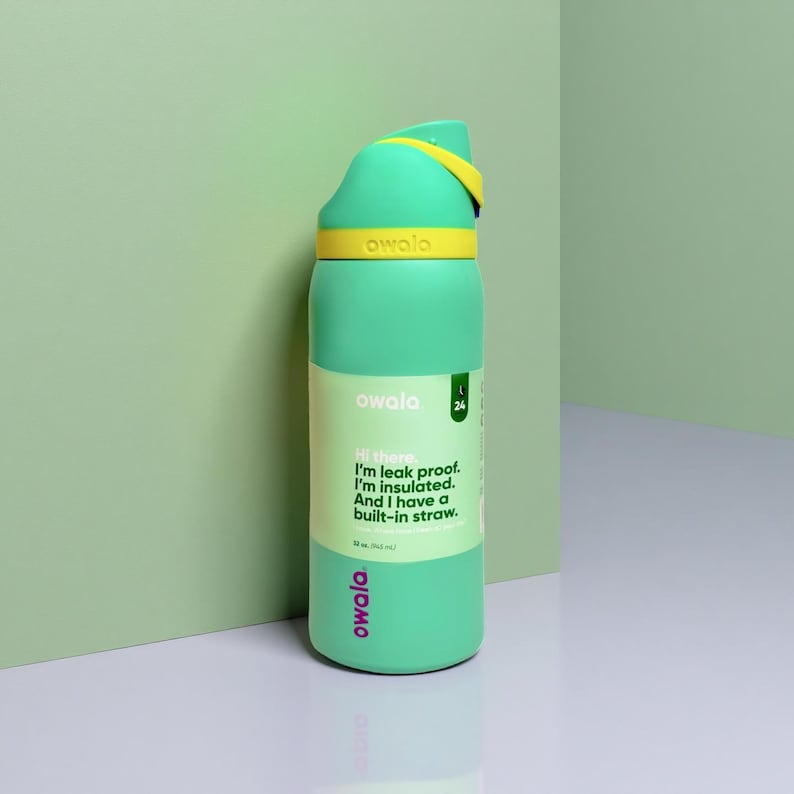 Puede incluir: Una botella de agua Owala verde menta con asa y detalles amarillos. La botella tiene el texto "owala" y "Hi there. I'm leak proof. I'm insulated. And I have a built-in straw."