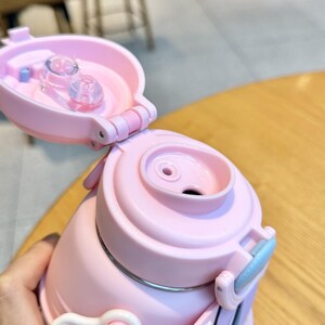 May include: Close-up of a pink insulated water bottle with a flip-top lid. The lid is open, revealing the drinking spout and a clear plastic component. The bottle has a light blue handle and a silver band around the middle.