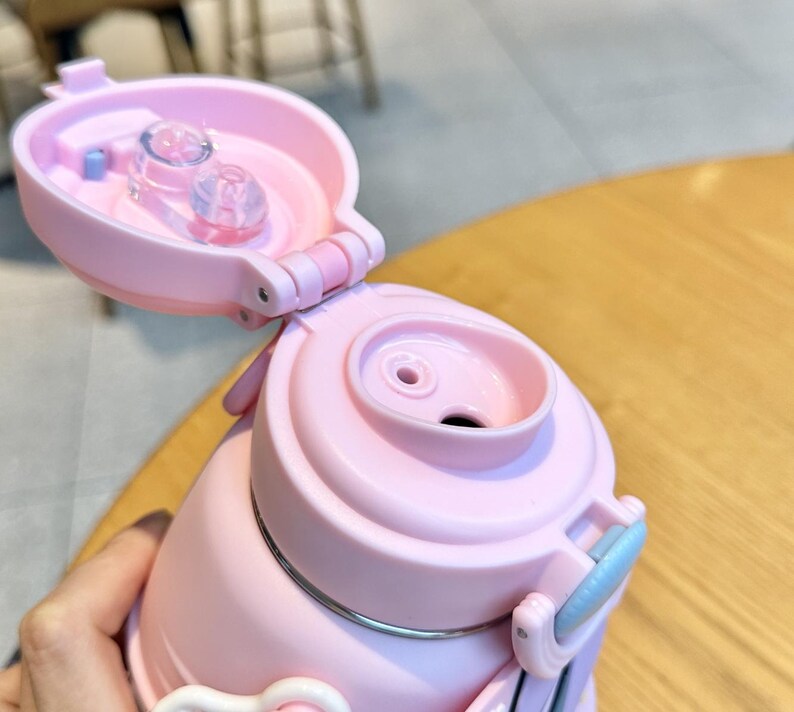 May include: Close-up of a pink insulated water bottle with a flip-top lid. The lid is open, revealing the drinking spout and a clear plastic component. The bottle has a light blue handle and a silver band around the middle.
