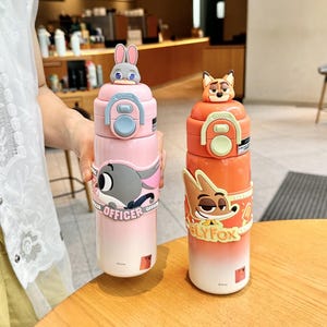以下が含まれることがあります： 2つの断熱ウォーターボトル。ピンクとオレンジで、漫画のキャラクターが描かれています。ピンクのボトルにはウサギのトッパーと「OFFICER」の文字が入ったウサギのグラフィックがあります。オレンジ色のボトルにはキツネのトッパーと「SLY FOX」の文字が入ったキツネのグラフィックがあります。