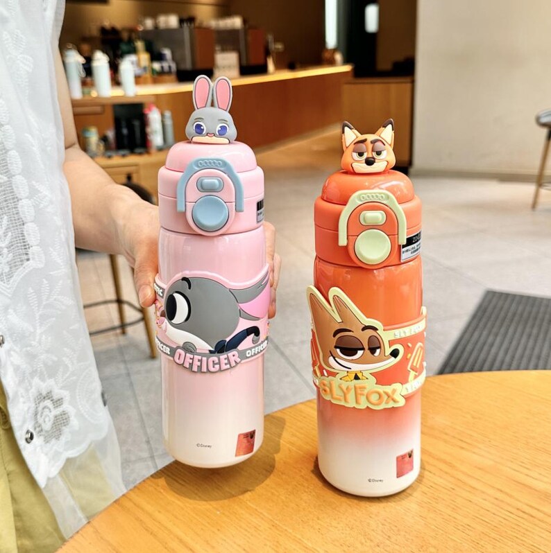 以下が含まれることがあります： 2つの断熱ウォーターボトル。ピンクとオレンジで、漫画のキャラクターが描かれています。ピンクのボトルにはウサギのトッパーと「OFFICER」の文字が入ったウサギのグラフィックがあります。オレンジ色のボトルにはキツネのトッパーと「SLY FOX」の文字が入ったキツネのグラフィックがあります。