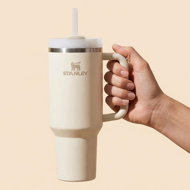 May include: Cream-coloured Stanley tumbler with a handle and a white straw. The tumbler has a silver band near the top and the Stanley logo. The tumbler is held by a hand.