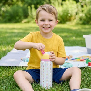 Puede incluir: Un ni&ntilde;o con una camiseta amarilla y pantalones cortos azules sentado en la hierba sostiene una botella de agua. La botella es blanca con un estampado floral y la palabra "awala". La botella tiene un asa y tapa rosas.