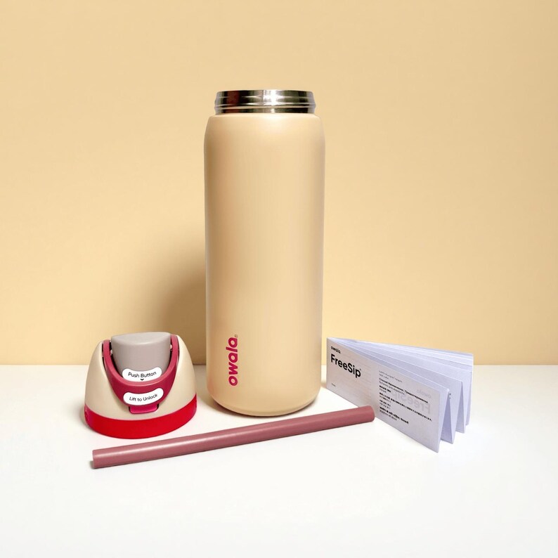 Op de afbeelding: Een beige Owala FreeSip ge&iuml;soleerde waterfles met een roestvrijstalen deksel, een rood en beige deksel, een roze rietje en een stapel witte instructiekaarten. De fles heeft het Owala-logo.