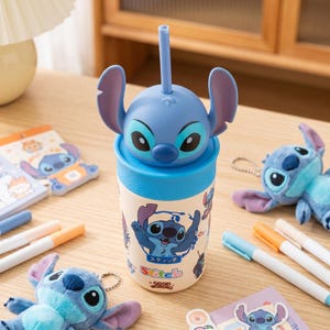 May include: A blue and white Stitch-themed tumbler with a straw, featuring a 3D character head on top. The tumbler has a cartoon graphic of Stitch and the text "Good Morning". Other Stitch-themed items, including a plush toy and notepads, are also visible.