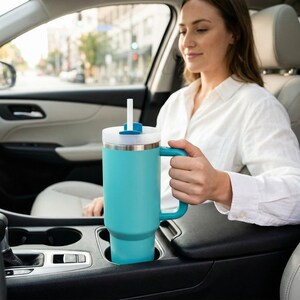 May include: A teal insulated tumbler with a handle and a white lid with a straw. The tumbler is in a car's cup holder. The person is holding the tumbler.