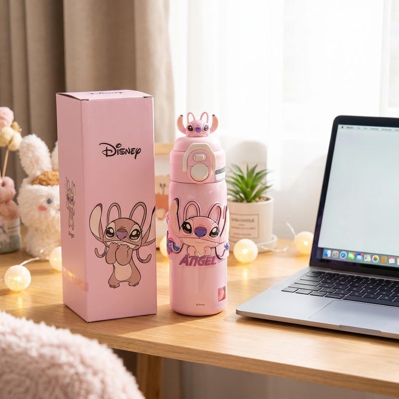 May include: A pink Disney water bottle with a cartoon character design, alongside a matching pink box. The bottle has a flip-top lid and a strap with the word "Angel". The box features the Disney logo.