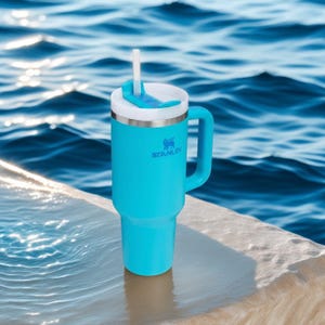 May include: A turquoise Stanley tumbler with a white lid and handle, featuring a straw. The tumbler has the Stanley logo and is set against a backdrop of water. The tumbler is designed to keep drinks cold.