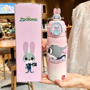 以下が含まれることがあります： ピンク色のズートピアをテーマにしたウォーターボトル。漫画のウサギのデザイン。ボトルは水色の蓋と、ズートピアのロゴとキャラクターのイラストが描かれたお揃いの箱が付いています。ボトルはピンクから白へのグラデーションでデザインされています。