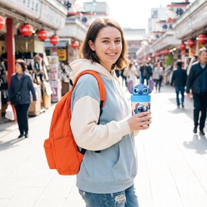 May include: A light blue and cream-colored hoodie with an orange backpack. A blue tumbler with a Stitch character design and a straw is held in the hand. The background shows a busy street with shops and people.
