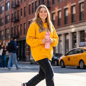 K&ouml;nnte beinhalten: Eine Person tr&auml;gt einen gelben Kapuzenpullover mit dem Wort "UNIVERSITY" in lila Buchstaben, eine schwarze Jeans und eine rosa Wasserflasche. Die Person geht auf einer Stadtstra&szlig;e mit einem gelben Taxi im Hintergrund.