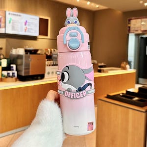 以下が含まれることがあります： ピンクと白のウォーターボトル。漫画のウサギとゾウのデザインが特徴です。 ボトルには、上部に小さなウサギのフィギュアが付いたフリップトップの蓋が付いています。 「OFFICER」の文字がボトルに印刷されています。 子供向けの楽しくて機能的なアイテムです。