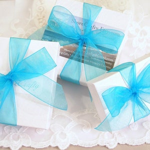 May include: Three white gift boxes tied with bright blue ribbon bows. The boxes are on a white lace surface. The top box has the text "Shore Crafty Frey.com" printed on it. The middle box has the text "Photo Charms, Wedding Gifts, Birthday Jewelry, and Groom Memory Charms" printed on it. The bottom box has the text "reCrafty@Gmail.com" printed on it.
