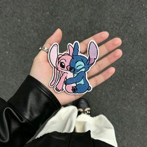 以下が含まれることがあります： 青いキャラクターとピンクのキャラクターが抱き合っている刺繍ワッペン。青いキャラクターは大きな耳と閉じた目を持ち、ピンクのキャラクターは長い耳と笑顔の表情をしています。ワッペンには白い縁取りと詳細なステッチが施されています。
