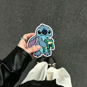 May include: An embroidered patch featuring a light blue alien character with large ears and a dark blue nose, holding a small green frog. The patch has a white border and is shaped to follow the outline of the characters in the design.