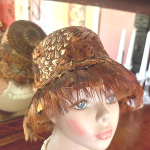May include: A cloche hat completely covered in layered brown, tan, and black feathers. The hat features a rounded crown and a short, downward-sloping brim. The feathers are arranged in a dense, overlapping pattern, creating a textured appearance.