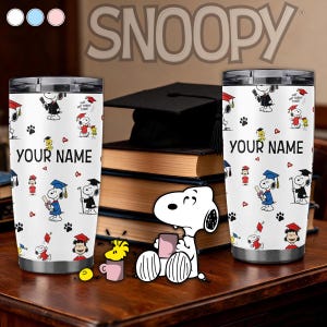 May include: Two white stainless steel tumblers with a graduation-themed Snoopy design. Each tumbler features the text "YOUR NAME" and illustrations of Snoopy in graduation attire. The background includes books and a graduation cap.