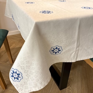 May include: White tablecloth with a repeating pattern of small, light gray flowers and larger blue floral designs. The tablecloth is draped over a table with dark legs, and the corner is visible.