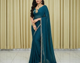 May include: A teal saree with gold trim and polka dots. The saree is draped over the shoulders and body. The person is wearing gold jewelry, including a necklace, earrings, bracelets, and rings. The background is a neutral color.