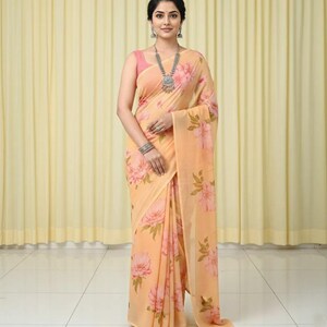 May include: A peach-colored saree with a large floral pattern in shades of pink and green. The saree is draped over a pink top. A silver necklace and earrings are worn.