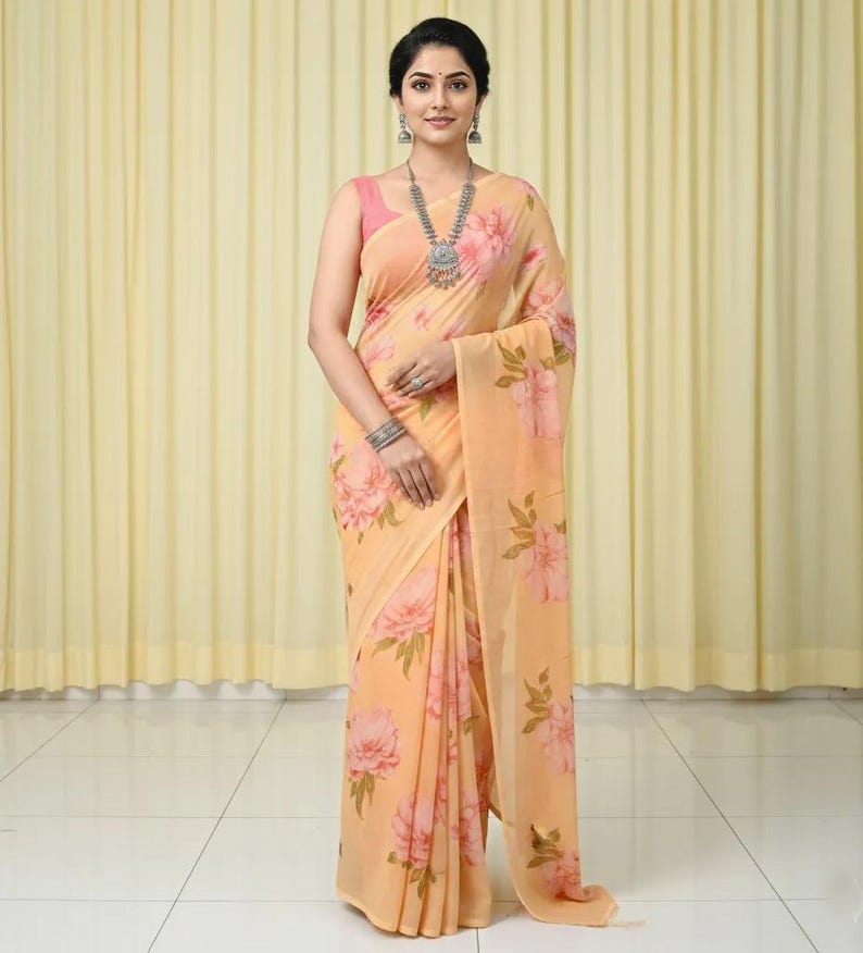 May include: A peach-colored saree with a large floral pattern in shades of pink and green. The saree is draped over a pink top. A silver necklace and earrings are worn.