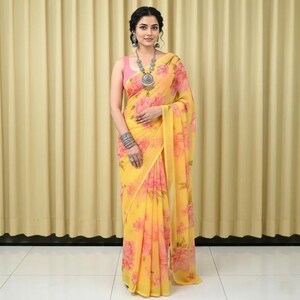 May include: A yellow floral saree with pink floral accents. The saree is draped over a pink top. The woman is wearing silver jewelry, including a necklace, earrings, and bracelets. The background is a light yellow curtain.