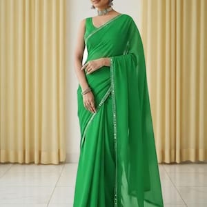 Puede incluir: Un sari verde esmeralda sin mangas con un borde brillante. La tela se drapea elegantemente, creando pliegues suaves. El sari es una prenda tradicional, a menudo usada para ocasiones especiales.