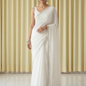 Puede incluir: Un sari blanco con una blusa sin mangas. El sari tiene un ribete plateado en los bordes. La tela cae elegantemente. El fondo es una cortina de color beige claro.