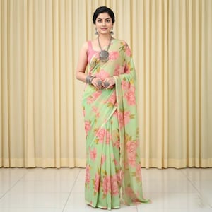 May include: A light green saree with a large pink floral pattern. The saree is draped over a pink top. The woman is wearing silver jewelry, including a necklace, earrings, and rings. The background is a light beige curtain.
