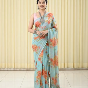 May include: A light blue saree with a floral pattern in shades of orange and peach. The saree is draped over a pink top. The woman is wearing silver jewelry, including a necklace, earrings, bracelets, and a ring.