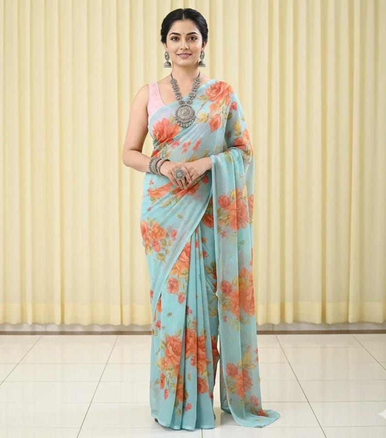 May include: A light blue saree with a floral pattern in shades of orange and peach. The saree is draped over a pink top. The woman is wearing silver jewelry, including a necklace, earrings, bracelets, and a ring.