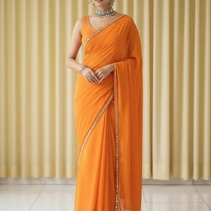 Puede incluir: Un sari naranja con una blusa sin mangas. El sari tiene un borde decorativo con peque&ntilde;os adornos. La mujer lleva un collar, pendientes y pulseras de plata. El fondo es una cortina de color amarillo claro.