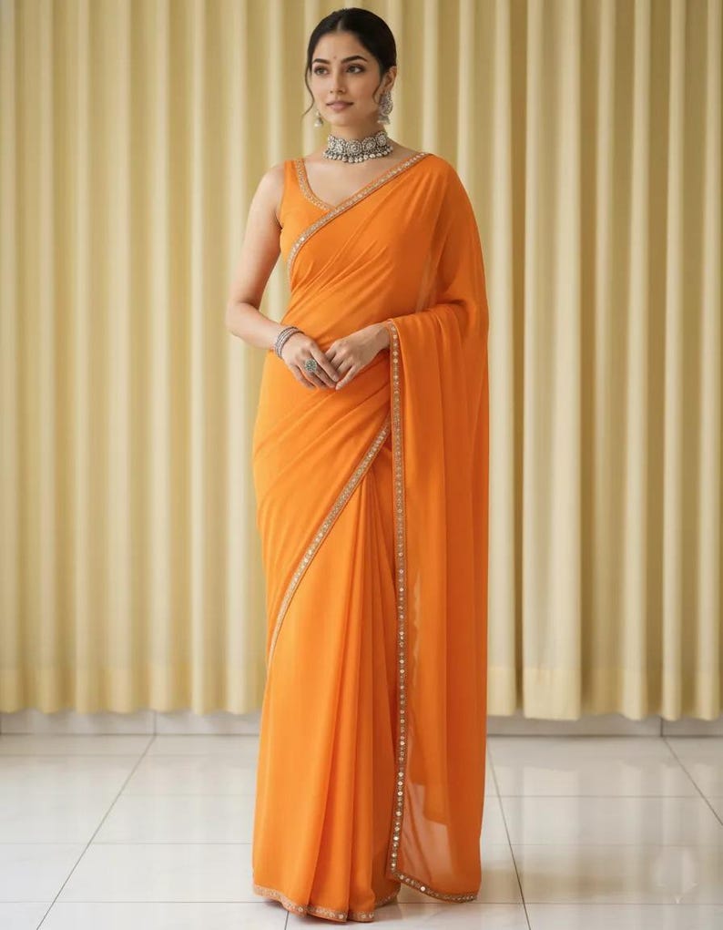 Puede incluir: Un sari naranja con una blusa sin mangas. El sari tiene un borde decorativo con peque&ntilde;os adornos. La mujer lleva un collar, pendientes y pulseras de plata. El fondo es una cortina de color amarillo claro.