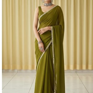 Puede incluir: Un sari verde oliva con un borde decorativo. El sari est&aacute; drapeado sobre el hombro y alrededor del cuerpo. La mujer lleva joyas, incluyendo un collar, pendientes y pulseras. El fondo es una cortina de color amarillo claro.