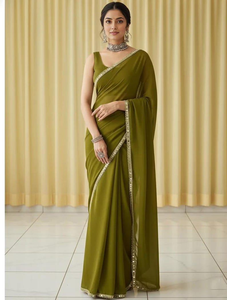Puede incluir: Un sari verde oliva con un borde decorativo. El sari est&aacute; drapeado sobre el hombro y alrededor del cuerpo. La mujer lleva joyas, incluyendo un collar, pendientes y pulseras. El fondo es una cortina de color amarillo claro.