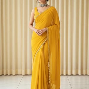 Puede incluir: Un sari amarillo vibrante con una blusa sin mangas. El sari presenta un delicado borde dorado y est&aacute; elegantemente drapeado. La mujer lleva joyas de plata, incluyendo un collar y pendientes. El fondo es beige neutro.