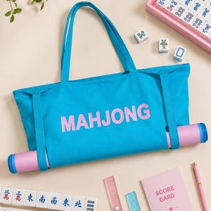Könnte beinhalten: Eine leuchtend blaue Tragetasche mit dem Wort MAHJONG in rosa Buchstaben auf der Vorderseite. Die Tasche hat zwei lange Tragegriffe und einen horizontalen Riemen an der Unterseite, der eine aufgerollte rosa-blaue Matte hält. Schlichtes Design.
