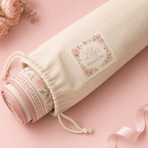 May include: A cream-colored drawstring storage bag containing a rolled mahjong mat. The bag features a square label with a floral wreath and the text Lily's Mahjong Mat. The mat inside has a pink border with a white Greek key pattern and floral motifs.