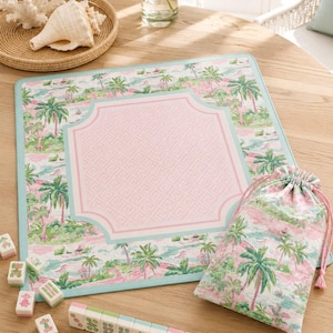 May include: A square mahjong mat featuring a tropical palm tree print in shades of pink and green with a light blue border and a central pink geometric frame. Includes a matching drawstring storage bag and a set of rectangular mahjong tiles.