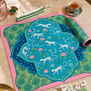 Puede incluir: Una alfombrilla de mahjong cuadrada con un panel central turquesa con leopardos blancos moteados y flores rosas, enmarcada por un borde festoneado azul oscuro y un patrón de agujas de pino verde, con un borde rosa sólido.