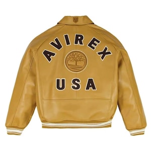 May include: A tan leather varsity jacket featuring a brown collar, ribbed cuffs, and hem with white stripes. The back displays the text AVIREX in an arc, a circular tree logo, and the text USA in large brown letters with white outlines.