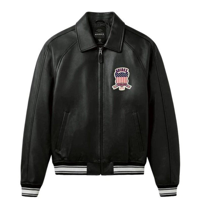 May include: Black leather jacket with a zip-up front, collar, and a patch on the chest. The jacket has white and black striped cuffs and hem. The patch features a design with the American flag and the word "AVIREX".