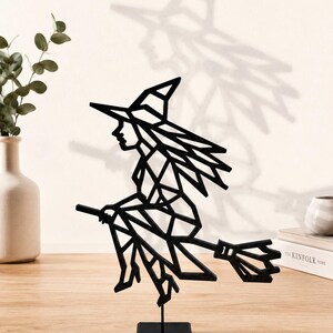 Op de afbeelding: Een zwarte geometrische heksenfiguur op een bezemsteel, gemonteerd op een kleine zwarte standaard. De heks draagt een puntmuts en heeft lang haar. Het beeldje staat op een houten oppervlak met een neutrale achtergrond.
